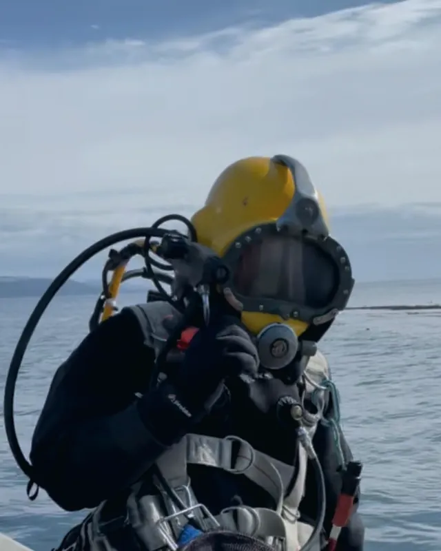 Commercial diver in a yellow KM helmet at the water's edge, giving the OK